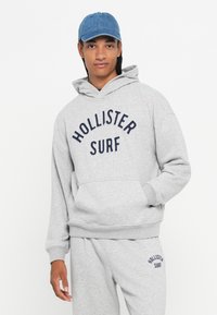 Mladic u sivoj dukserici "Hollister Surf" i trenirkama, s plavom traper kapom, stoji ispred bijele pozadine.