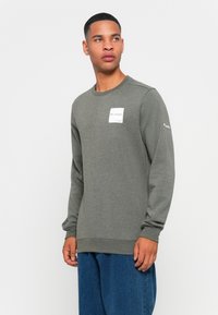 Mann iført en grå Columbia hettegenser med logoer på brystet og armen, sammen med løse blå jeans, står mot en enkel hvit bakgrunn.