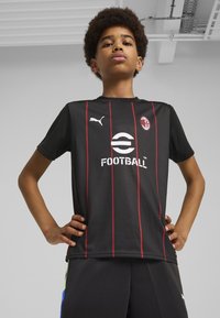 Zwart voetbalshirt met rode verticale strepen, voorzien van een Puma-logo, ronde halslijn en centrale "FOOTBALL" print. Draagt donkergekleurde shorts.