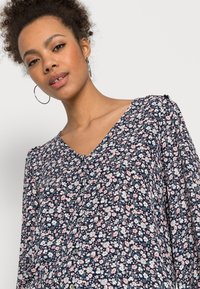 Bluză florală de culoare albastru închis cu flori roz și albe, decolteu în V, mâneci lungi și detalii cu nasturi pe față. Material ușor.