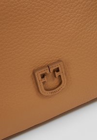 Gros plan sur un cuir tanné texturé avec un patch logo Furla cousu et en relief de couleur assortie.