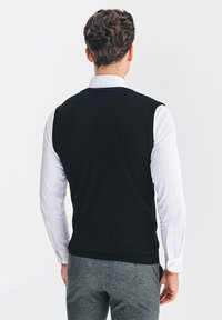 Czarny bezrękawnik z prążkowanym wykończeniem, noszony na białej koszuli, w parze z szarymi spodniami. Gładka tekstura, casualowy krój.