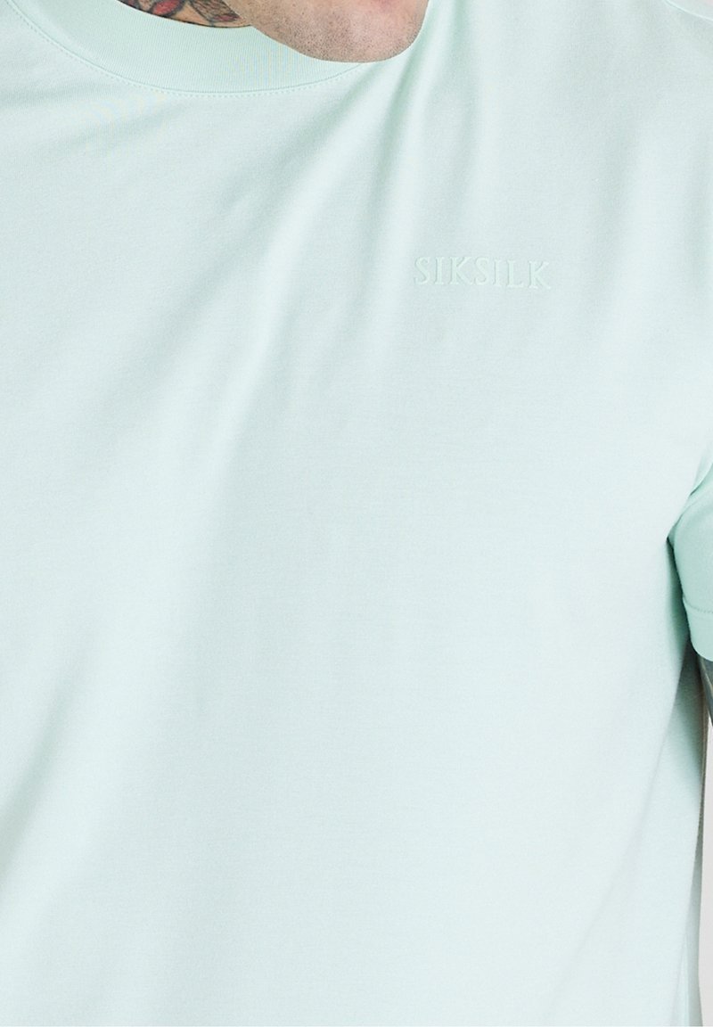 Světle mátově zelené tričko s krátkým rukávem vyrobené z měkké látky, s texturovaným logem vyšitým uprostřed. Jednoduchý, přiléhavý design.