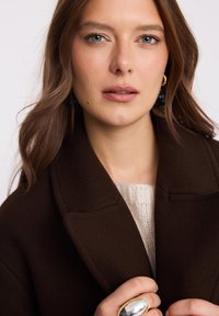 Brązowy wełniany płaszcz z wyłamią kołnierza, trzymany zamknięty ręką. Pod nim widoczny jest jasny sweter z dzianiny. Duży okrągły pierścień na ręce.