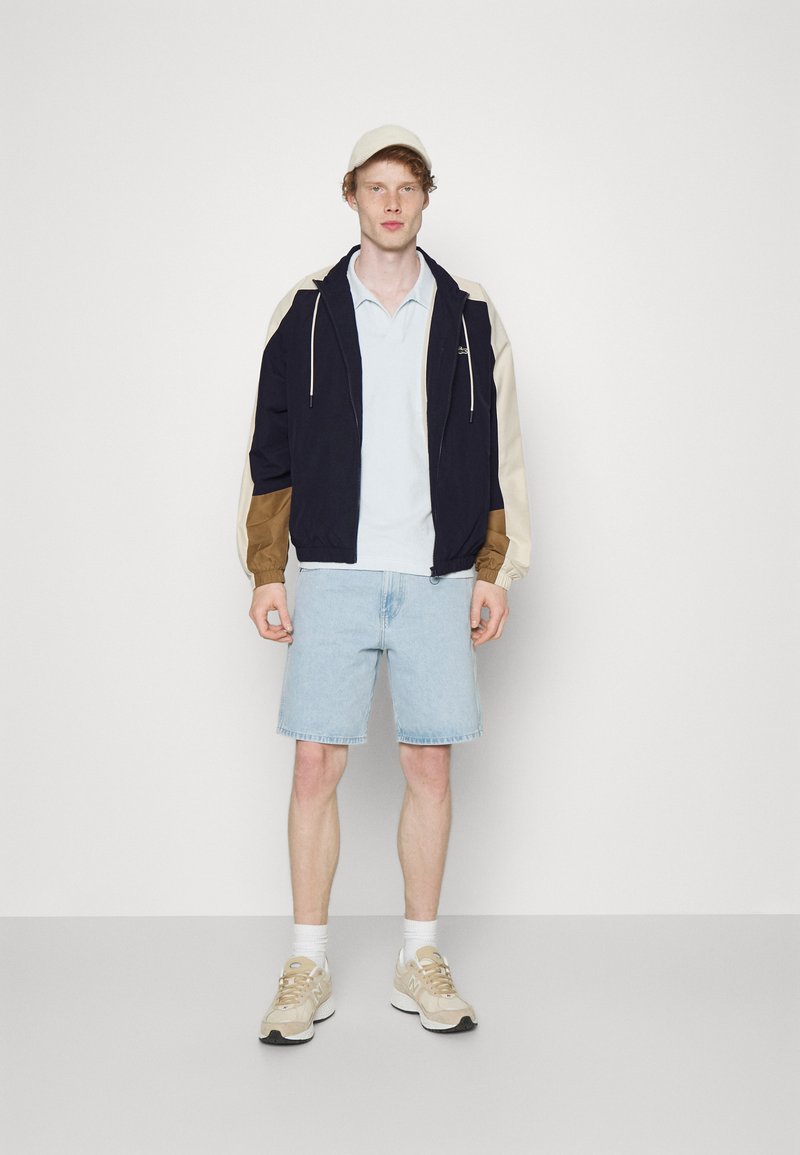 Marineblauw en beige kleurblokjack met capuchon en koorden; lichtblauwe polo; denimshorts; tan sneakers met witte sokken.