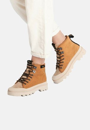 Botas altas de ante color tan con suela de goma texturizada beige, que presentan ojales de metal y cordones marrones. Acento negro en el cuello.