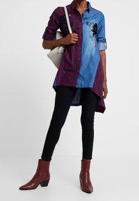 Donna che indossa una tunica asimmetrica con un design metà a quadri e metà in denim, pantaloni skinny neri, stivaletti borgogna e una borsa a tracolla grigia chiara.