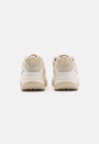 Beige sneakers med vadderad häl, texturerade mockadetaljer och vit mellansula. Har en dragloophank baktill och en robust sula.