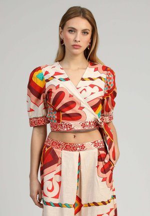 Femme portant un cache-cœur court à motifs colorés avec des manches bouffantes et une jupe taille haute assortie, debout devant un fond uni.