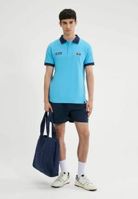 Polo shirt azzurro chiaro con dettagli blu navy, etichetta del logo, abbinato a pantaloni corti blu navy e una borsa tote blu navy. Il modello indossa sneakers bianche e calzini alla caviglia.