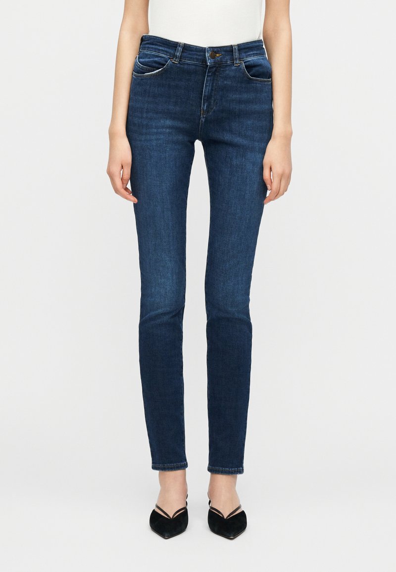 Στενά τζιν σε σκούρο μπλε χρώμα σε όρθιο άτομο που φοράει μαύρα παπούτσια slingback με μυτερή μύτη και λευκό τοπ, σε απλό φόντο.