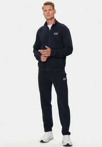 Uomo che indossa un completo EA7 nero con sneakers bianche, in piedi con le mani incrociate contro uno sfondo bianco uniforme.