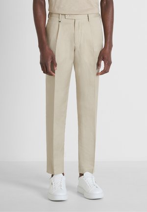 Homme portant un pantalon beige sur mesure et des baskets blanches, se tenant devant un fond clair et uni.