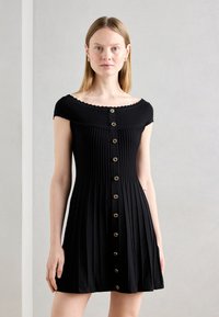 Robe noire côtelée avec un décolleté ondulé à épaule dénudée, boutons sur le devant et une jupe évasée. Tissu lisse et texturé.