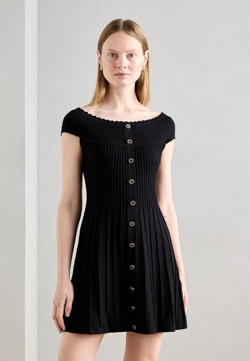 Robe noire côtelée avec un décolleté ondulé à épaule dénudée, boutons sur le devant et une jupe évasée. Tissu lisse et texturé.