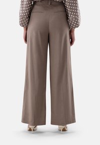 Weite, braune Hose aus leichtem Stoff. Merkmale sind ein sauberer Schnitt, zwei Gesäßtaschen und eine glatte Textur. Kombiniert mit beigen Absatzschuhen.