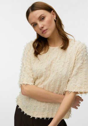 Jeune femme aux cheveux châtain clair porte un chemisier crème texturé à manches courtes et de petites boucles d'oreilles anneaux, posant les bras croisés.