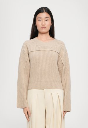 Beige ribbadus med bred rund hals, långa ärmar och texturerad design, i kombination med ljusa, veckade byxor.