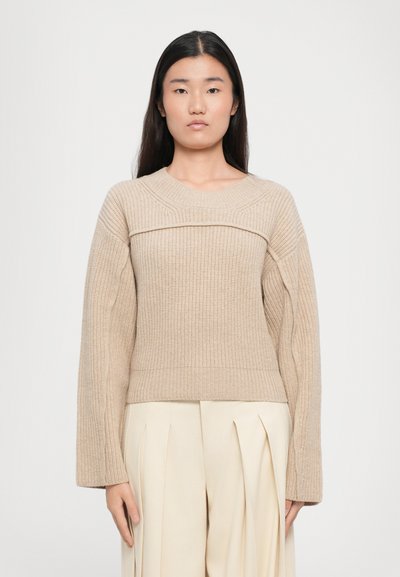 Beige gerippter Pullover mit weitem Rundhalsausschnitt, langen Ärmeln und strukturiertem Design, kombiniert mit hellen, gefalteten Hosen.