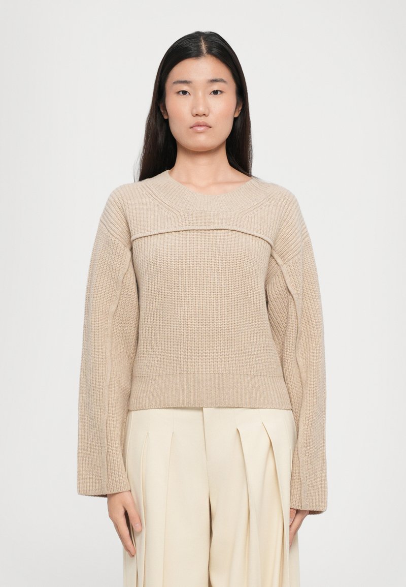 Maglione beige a coste con ampio collo girocollo, maniche lunghe e design texturizzato, abbinato a pantaloni plissettati di colore chiaro.