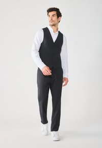 Gilet noir sur une chemise blanche, associé à un pantalon noir et des baskets blanches. Tissu lisse avec une coupe ajustée et cinq boutons.