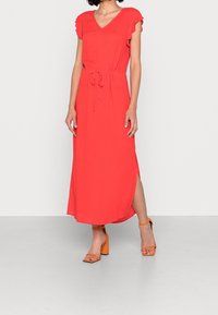 Robe rouge longueur genou en tissu léger, dotée d'un col en V, de manches volantées, d'une taille ajustable avec cordon et de fentes latérales. Associée à des talons carrés orange.