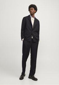Abito blu navy con giacca a un bottone e pantaloni coordinati, indossato sopra un maglione leggero e una camicia a righe, completato da scarpe nere.