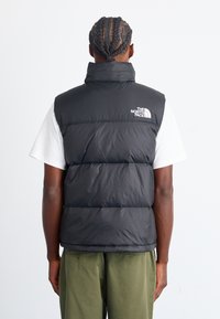 Black puffer vest with high collar and branded logo on the back. Features quilted design, shiny texture, and front zipper closure.