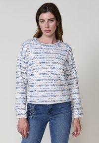 Pull en maille doux et texturé avec des rayures bleues et beiges. Il dispose d'un col bateau et de manches longues. Porté avec un jean en denim brodé.