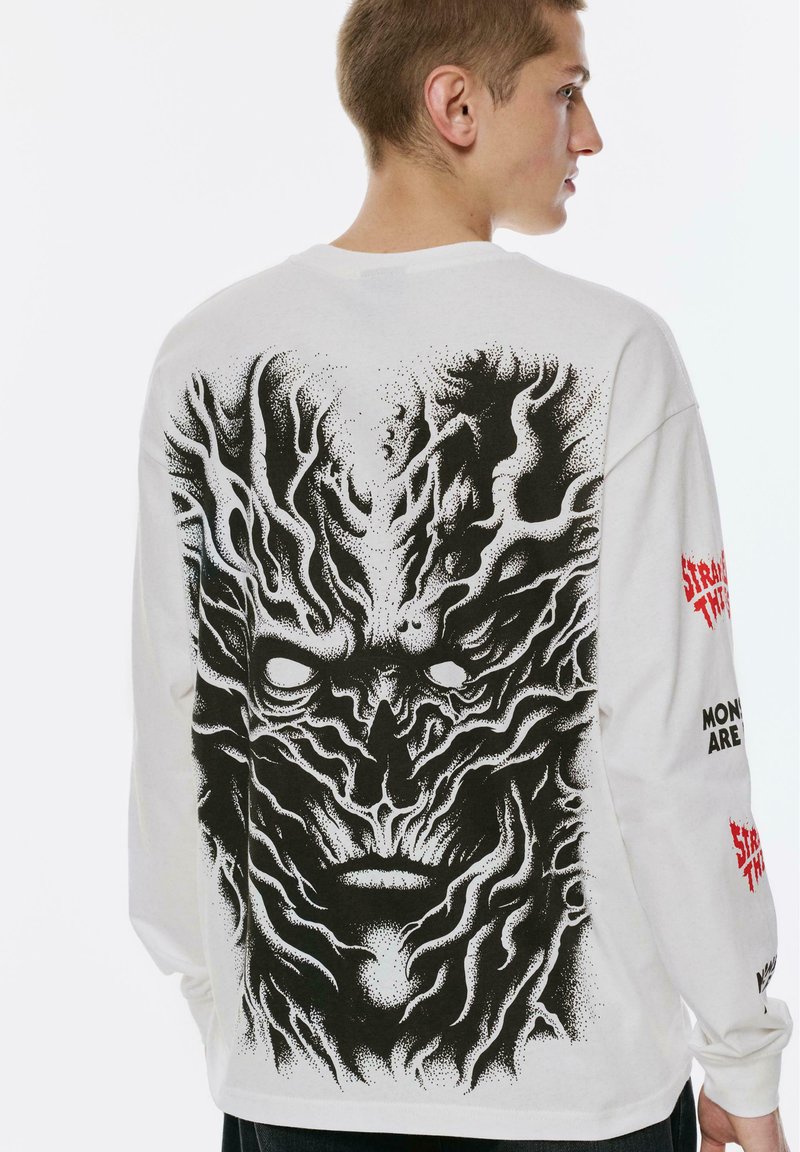 Junger Mann in einem weißen Langarmshirt mit großem, schwarzem, abstraktem baumähnlichem Gesichtsdruck auf dem Rücken und rotem Text auf den Ärmeln.