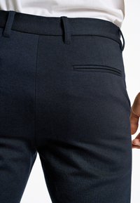 Donkerblauwe broek van geweven stof. Kenmerken zijn een platte voorkant, riemlussen, slim fit en een enkele achterzak met een slank ontwerp.