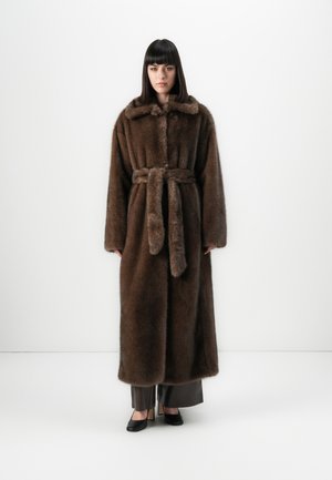 Donna con capelli neri lisci, che indossa un lungo cappotto in pelliccia sintetica marrone con cintura, pantaloni larghi e tacchi neri, in piedi di fronte a uno sfondo bianco.