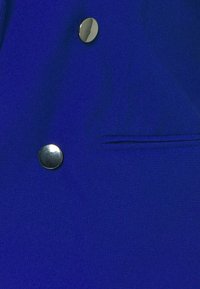 Blauwe stoffen blazer met een gladde textuur, voorzien van zilveren knopen en een klein zakje aan de linkerkant voor extra detail.