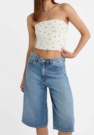 Mujer con un top corto blanco floral sin tirantes y pantalones cortos de mezclilla azul claro sueltos, con un collar en forma de corazón y mano en la cadera.