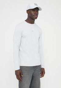 Helehall pikkade varrukatega särk, millel on ümmargune kaelus ja peenike "BOSS" logo keskel. Koos hallide teksade ja valge nokamütsiga.