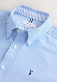 Hellblaues Hemd mit Knöpfen, weißen Knöpfen, gestreiftem innerem Kragen und kleinem dunkelrotem Hirsch-Logo auf der Brust, Größe Medium von der Marke NEXT.