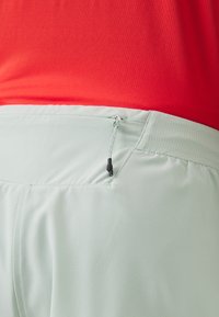 Bermudas deportivas de color verde claro con una cinturilla texturizada y un cordón negro, combinadas con una parte superior roja. Primer plano del detalle del bolsillo trasero.