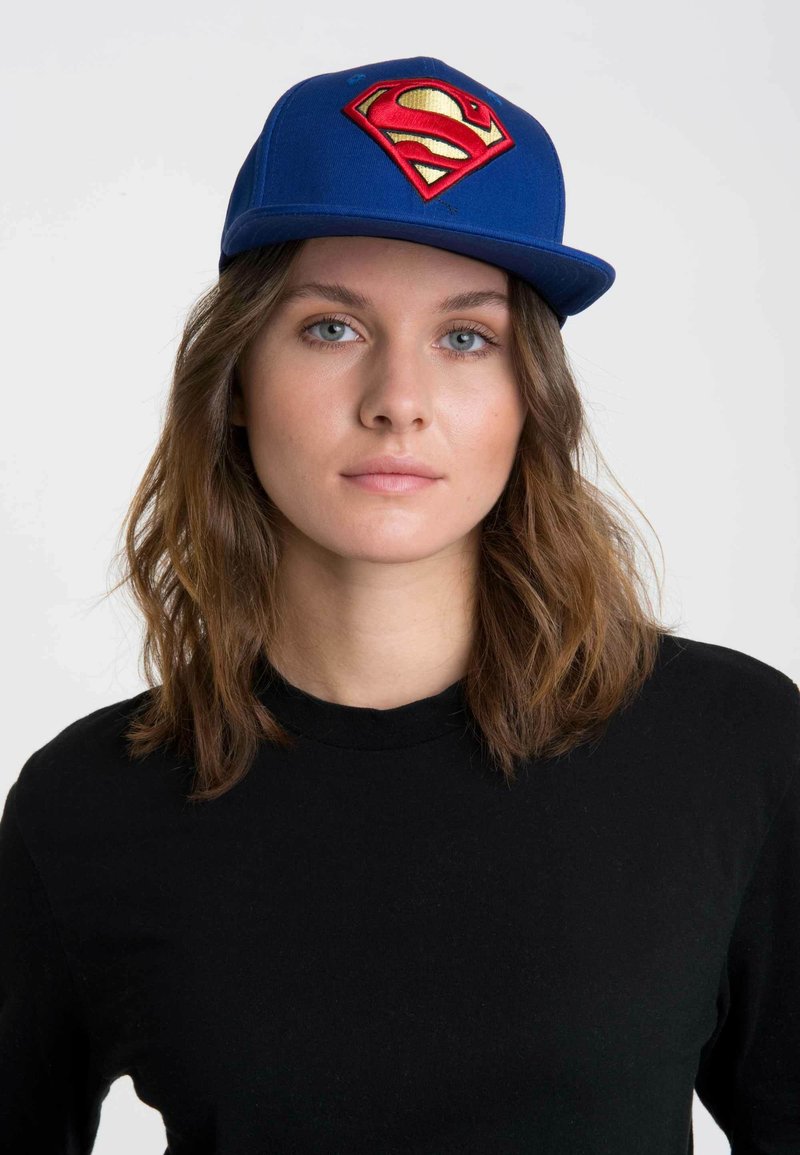Gorra azul con visera plana y un logotipo bordado en rojo y dorado de Superman en la parte frontal. Se lleva con una camiseta de manga larga negra.