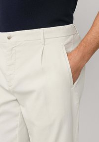 Muškarac u bež hlačama s jednom rukom u džepu, uparen s crnom majicom, prikazan od struka do sredine bedra na neutralnoj pozadini.