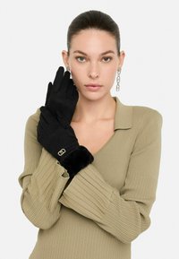 Guantes negros de material suave, con un puño de piel de pelo y un acento de logo dorado. Modelo vistiendo una blusa de color oliva con canalé.