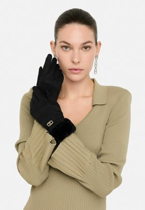 Guanti neri realizzati in materiale morbido, con polsino in pelliccia e un'elegante accentuazione in oro. Modella che indossa un top rigato color oliva.