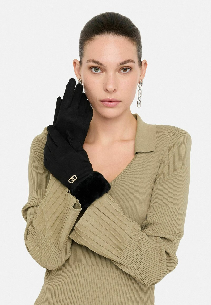 Guantes negros de material suave, con un puño de piel de pelo y un acento de logo dorado. Modelo vistiendo una blusa de color oliva con canalé.