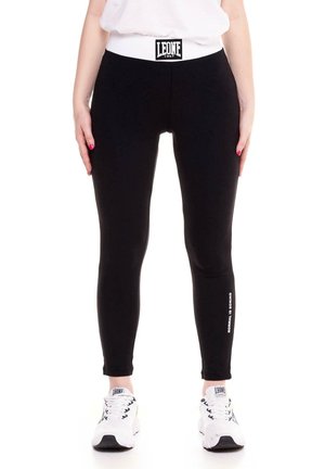 Modèle portant un legging de sport noir long, avec une ceinture blanche Leone et une inscription sur le côté, associé à des baskets blanches.
