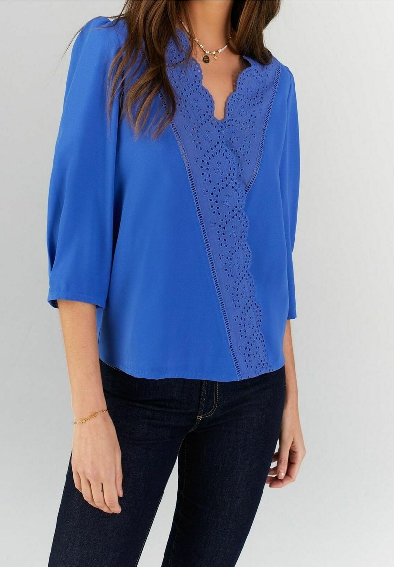 Blaue Bluse mit V-Ausschnitt, kurzen Ärmeln und filigraner Lochmusterung auf einer Seite. Der Stoff ist glatt und leicht.