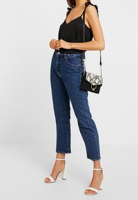 Haut noir sans manches avec bretelles nouées ; jean bleu taille haute ; talons blancs à lanières ; sac bandoulière effet peau de serpent noir et blanc avec bandoulière chaîne.