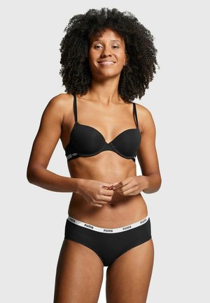 Woman with curly hair wearing black bra and black Puma briefs with white waistband, standing against light gray background.