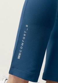 Siniset stretch-kankaasta valmistetut capri-housut, joissa on sileä tekstuuri ja mukana näkyvä, vaaleanharmaa "COMFORT_B" -logo sivussa.