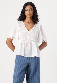 Blusa bianca con scollatura a V, maniche corte a sbuffo e dettagli in pizzo. Presenta una chiusura con bottoni e una vestibilità rilassata. Tessuto leggero e testurizzato.