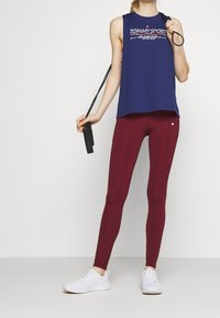 Top sportivo senza maniche di colore blu navy con il logo "TOMMY SPORT", abbinato a leggings borgogna. La modella tiene una corda per saltare e indossa sneakers bianche.
