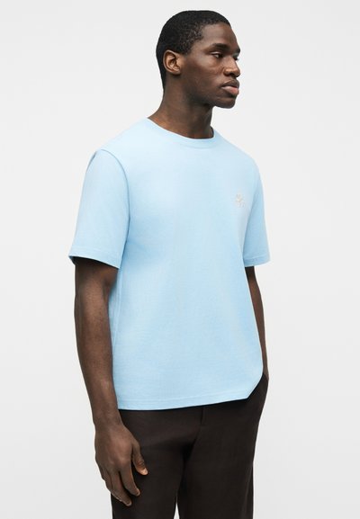 Homme debout, les mains dans les poches, portant un t-shirt bleu clair à manches courtes et un pantalon sombre, devant un fond blanc uni.